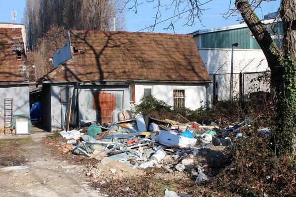 前にごみの山と背景に澄んだ青空に囲まれたワイヤーフェンスで区切られた産業団地の隣にある背の高い木の陰の小さな家族の家 — ストック写真