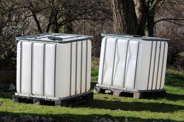 Brand new two intermediate bulk containers or IBC plastic tanks with metal cage used for water storage put on wooden pallets in local garden surrounded with grass and trees on warm sunny spring day clipart