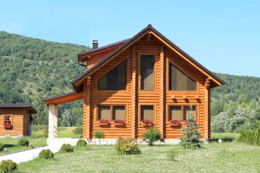 Arka planda açık mavi gökyüzü ile küçük çiçekler ve orman dolu kesilmemiş çim ile çevrili büyük pencereler ve taş girişi ile yeni inşa ahşap günlük kabin ev