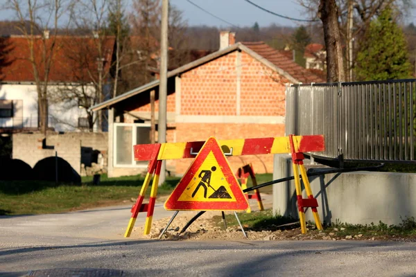 Work ahead road sign at the side of paved road next to concrete and metal fence with red brick family houses in background on warm sunny spring day