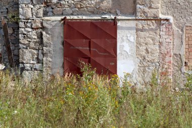 Paslanmış metal sürgülü ambar kapıları uzun boylu çim ve sıcak güneşli günde terkedilmiş sanayi kompleksi önünde küçük çiçekler ile eski harap duvara monte