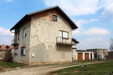 Suburban family house heavily damaged by shrapnel during war with dilapidated facade, rusted garage doors and balcony handrails, visible bricks, new windows and blinds surrounded with winter grass, gravel and cloudy sky in background clipart