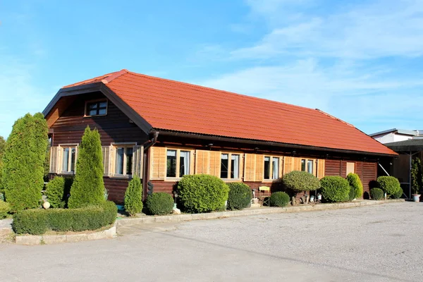 Elongated ground level wooden restaurant with new roof tiles surrounded with gravel parking and various shapes of hedges on cloudy blue sky background