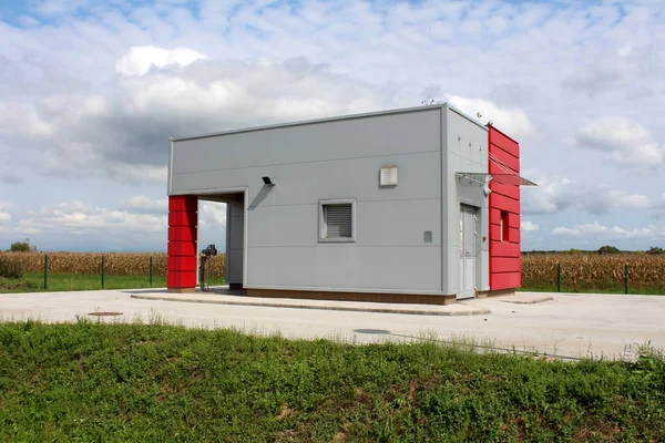 Industrial water pumping station inside concrete structure with red and grey metal walls surrounded with asphalt and grass with cornfield and cloudy blue sky in background