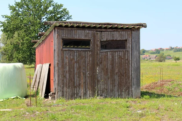 Old dilapidated wooden garage with two doors and missing door handle now used as garden shed for storing tools surrounded with uncut grass mixed with small flowers and large tree with family houses in background on warm summer day