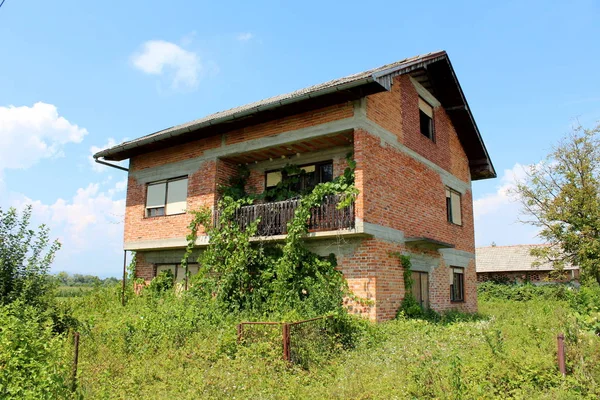 Red bricks abandoned family house with broken windows and front wire fence overgrown with high uncut grass, trees and crawler plants with backyard building and cloudy blue sky in background on warm sunny day