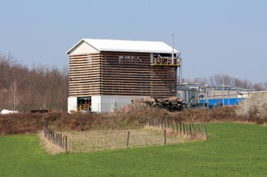 Ahşap tahtalar ve metal çatı dan yapılmış duvarlar ile büyük sanayi binası önünde taze çim ve arka planda açık mavi gökyüzü ile endüstriyel kompleks ve kuru orman bitki örtüsü ile çevrili