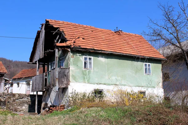 Abandoned old family house with broken roof tiles and cracked front wooden porch disconnected from power grid with hanging electrical wires surrounded with uncut grass and other houses in background on warm sunny spring day