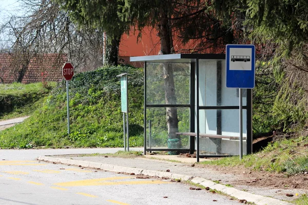 Glass covered transparent local bus station with wooden bench next to traffic signs and notice board surrounded with uncut grass and paved road on warm sunny spring day