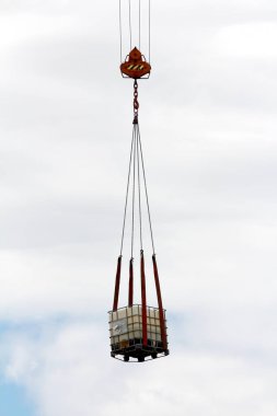 İnşaat işçilerine su getirmek için kullanılan metal kafesli ara dökme konteyner veya Ibc plastik tankı, bulutlu gökyüzü arka planında inşaat alanının üzerinde ağır inşaat vinci ve dört güçlü zincirle havada yüksekten kaldırıldı