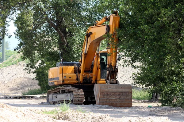 暖かい晴れた春の日に大きな建設現場でトラックが積み込まれるのを待っている大きな木陰に駐車した掘削機 — ストック写真