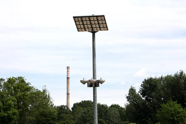 LED modern street light reflectors pointed towards large reflective panels mounted on top of strong metal pole with industrial chimney in background surrounded with dense trees on warm sunny spring day