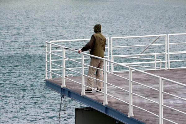 Man in hunting jacket and hoodie standing at the end of local pier fishing with small fishing rod over white metal fence on rainy spring day