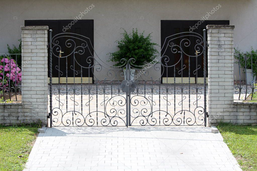 Large wrought iron family house driveway entrance doors mounted on two stone tiles covered concrete poles leading towards two garage doors on warm sunny summer day