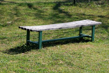 Sıcak güneşli bahar gününde yerel parkta taze kesilmiş çim ile çevrili ahşap koltuk ve yeşil metal destek çerçevesi ile eski kamu park bankı