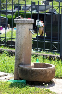 Sıcak güneşli yaz gününde taş fayans ve kesilmemiş çim ile çevrili mezarlık metal çit önünde parlak metal musluk ile Beton kamu su sebili