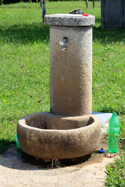 Sıcak güneşli yaz gününde açık plastik şişe ve kesilmemiş çim ile çevrili beton temel üzerine inşa edilmiş parlak metal musluk ile beton kamu su sebili ön görünümü