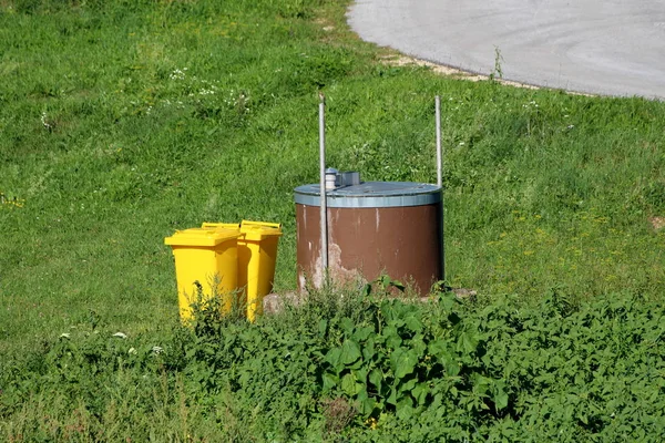自制棕色的井 上锁有闪亮的金属盖 旁边是两个明亮的黄色塑料垃圾桶 周围是未切割的草和铺面的道路 在温暖的阳光明媚的夏日 — 图库照片