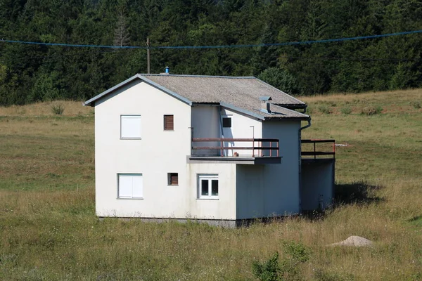 Large abandoned family house with closed window blinds surrounded with uncut grass and dense forest in background on warm sunny summer day