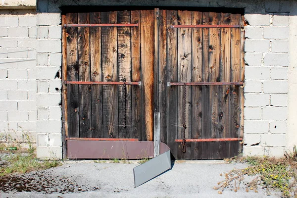 Old wooden garage doors with faded wooden boards and broken metal protection mounted on abandoned outdoor storage building surrounded with paved driveway partially covered with grass and moss on warm sunny summer day
