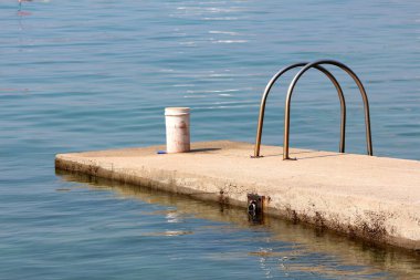 Daha kolay giriş için kısmen paslanmış metal tırabzanları olan beton iskele ve sıcak yaz gününde sakin, açık mavi denizle çevrili küçük tekneleri bağlamak için metal kanca.