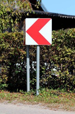 Yeşil çitli ve arka planda evi olan asfaltlı yolun yakınındaki metal direğe asılmış tehlikeli dönüş kırmızı ve beyaz ok işareti.