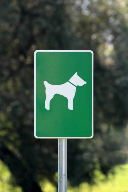 Üzerinde beyaz köpek olan yeşil metal tabela köpeklerin arka planda ağaçlarla birlikte gri direğe monte edilmiş bu parka girmelerine izin verildiğini gösteriyor.