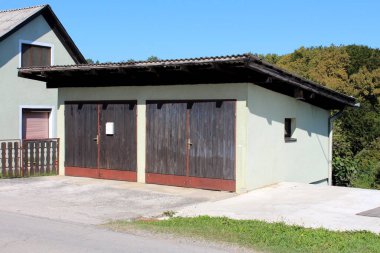 Açık yeşil açık hava garajı. Küçük tepenin kenarındaki iki ahşap kapı. Yoğun orman ve arka planda açık mavi gökyüzü ile çevrili bir aile evi ve beton yol.