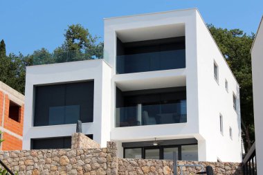 Beyaz cepheli, camlı balkon çitli, siyah pencereli, geleneksel taş duvarlı ve arka planda güneşli yaz gününde ağaçlarla çevrili modern bir apartman.