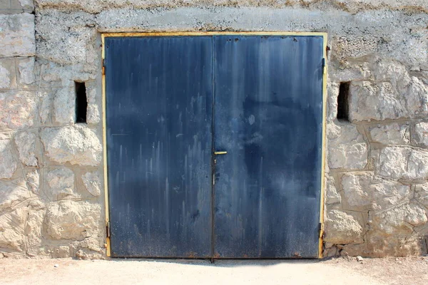 Dark blue dilapidated metal garage doors with rusted yellow frame mounted on stone wall with two openings for ventilation next to paved road on warm sunny summer day