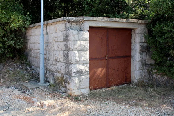 Detached small garage with rusted metal doors and stone walls next to gravel road surrounded with dense trees and vegetation on warm sunny summer day