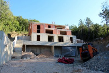 Tamamlanmamış büyük kırmızı bina, açık mavi arka planda inşaat malzemesi ve yoğun ağaçlarla çevrili küçük tepenin kenarındaki aile inşaat alanını engelliyor.