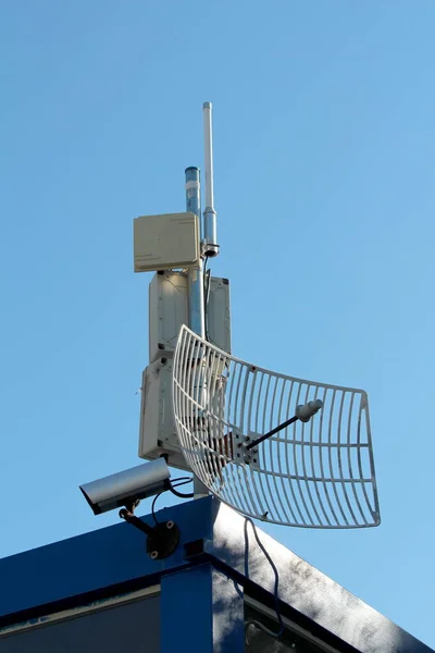 TV antennas with transmitters and closed circuit TV CCTV security camera densely mounted on single pole on top of industrial complex building on clear blue sky background