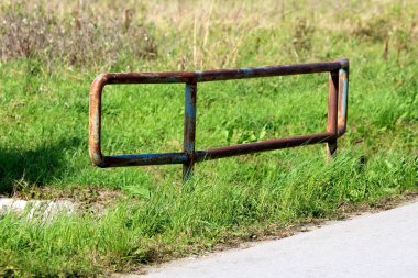 Küçük asfalt köprünün kenarında paslanmış güçlü metal koruma çitleri. Sıcak bir sonbahar gününde etrafı kesilmemiş çimlerle çevrili.