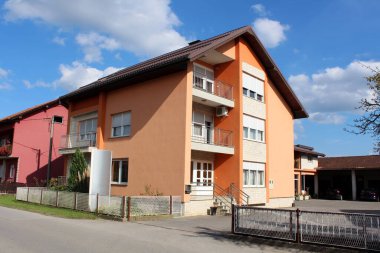 Yeni inşa edilmiş turuncu banliyö aile evi geniş asfaltlı ön bahçesi ve metal çitlerle çevrili iki açık garaj ve bulutlu mavi gökyüzü arka planında uzamış metal kapılar.