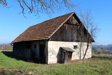 Ahşap duvarları olan harap olmuş terk edilmiş küçük bir ahır ve açık mavi gökyüzü arka planında çimen ve ağaçlarla kaplı küçük bir tepenin tepesinde çatlamış beton bir temele yaslanmış bir tuvalet.