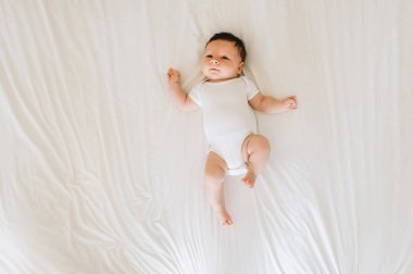 overhead view of cute newborn baby in white bodysuit lying on bed clipart