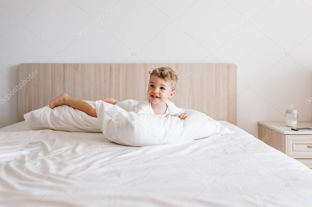 smiling toddler boy in white bodysuit lying on pillows on bed at home