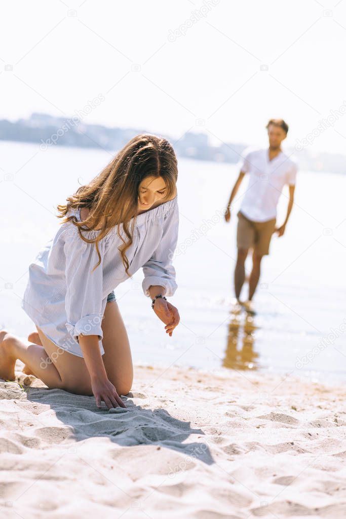 girlfriend sitting on city beach near river and playing with sand