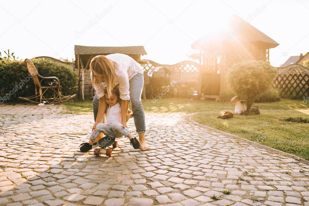 mother helping little daughter skateboarding on backyard on summer day