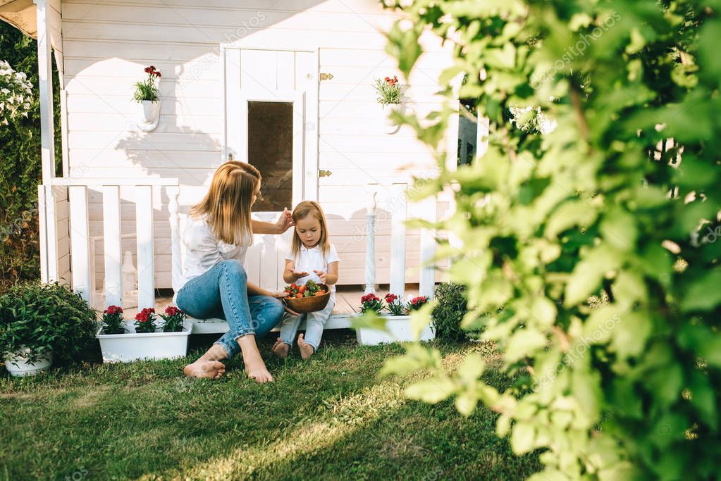 Mother and adorable little kid with bowl of strawberries resting on porch of little country house together