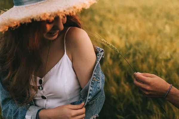 Attractive smiling girl in straw hat sitting on meadow, while boyfriend touching with wheat ear to her shoulder — Stock Image Attractive Smiling Girl Straw Hat Sitting Meadow While Boyfriend Touching Royalty Free Stock Images