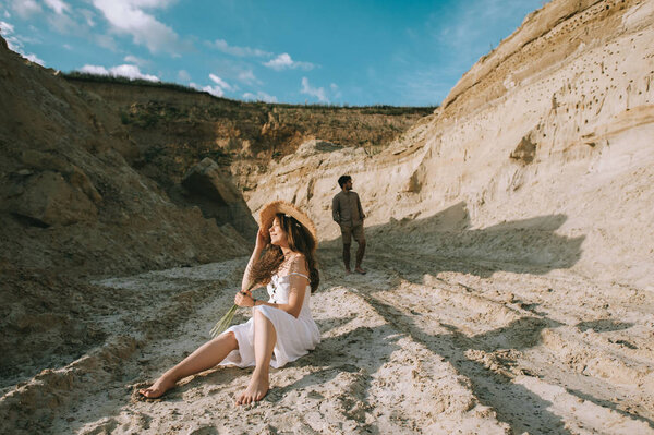 счастливая девушка с цветочным букетом сидит в песчаном каньоне с парнем позади
