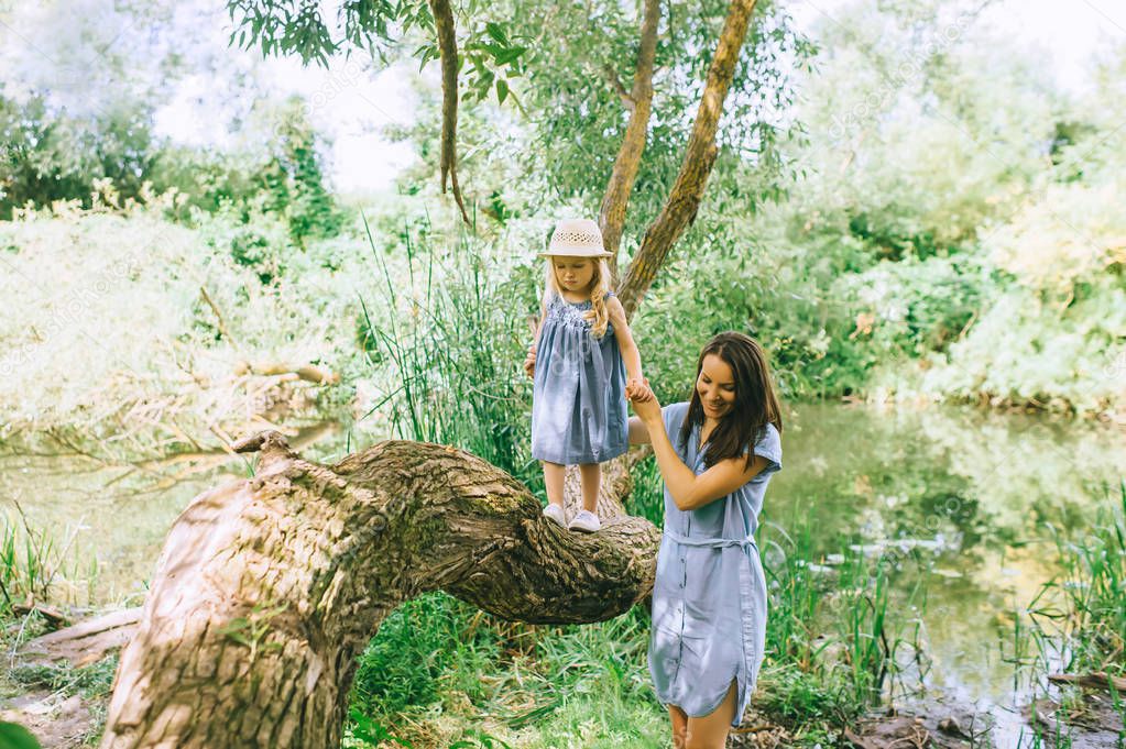mother holding hands with daughter and helping her to walk on tree trunk, with lake on background