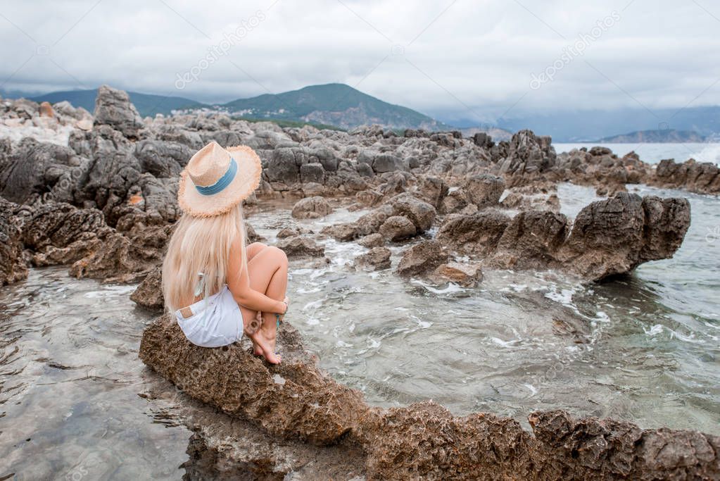 girl in bikini top and straw hat sitting on rocky beach in montenegro