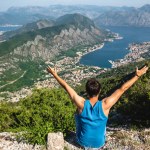 Back view of man sitting with raised hands and looking at Kotor bay and Kotor town in Montenegro