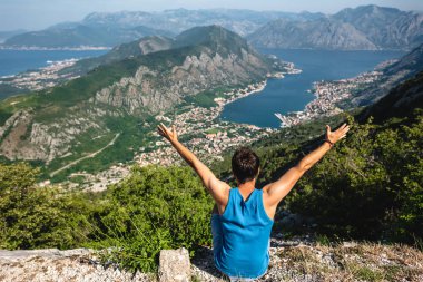 back view of man sitting with raised hands and looking at Kotor bay and Kotor town in Montenegro clipart
