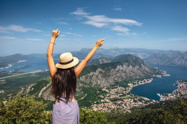 back view of woman standing with raised hands and looking at Kotor bay and Kotor town in Montenegro clipart