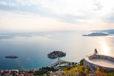 woman sitting on viewpoint and looking at island of Sveti Stefan during sunset in Budva, Montenegro clipart