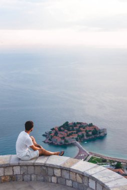 man sitting on viewpoint and looking at island of Sveti Stefan with hotel resort in Adriatic sea, Budva, Montenegro clipart